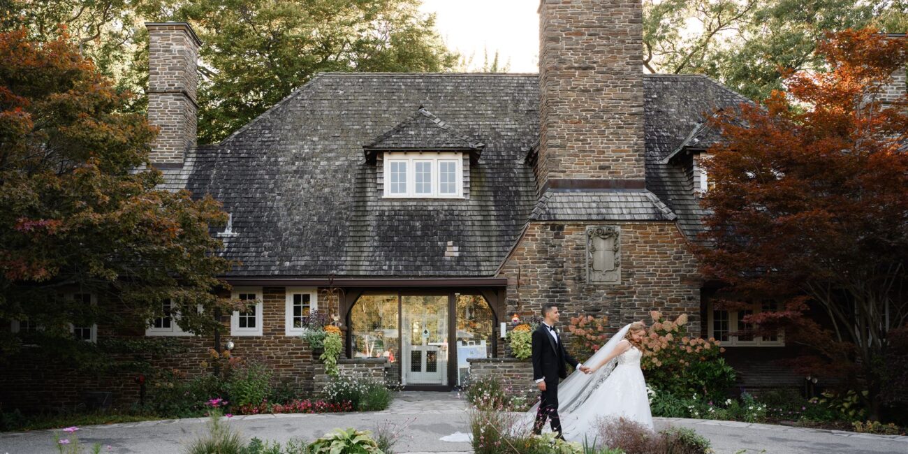 A couple in wedding attire poses in front of a large stone house surrounded by trees and a small garden, embodying the charm of Oakville wedding venues.