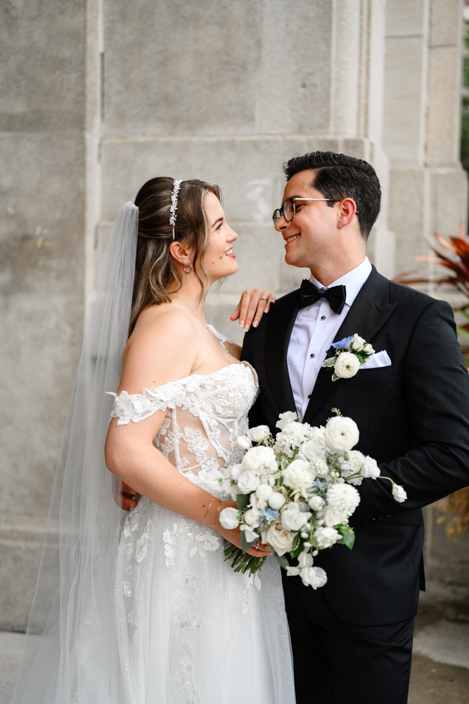 The bride and groom share a loving glance outside, holding a bouquet of white flowers. She is elegant in a laced white gown, and he looks dapper in a black tuxedo with a bow tie, capturing the magic of their Liberty Grand Toronto wedding.