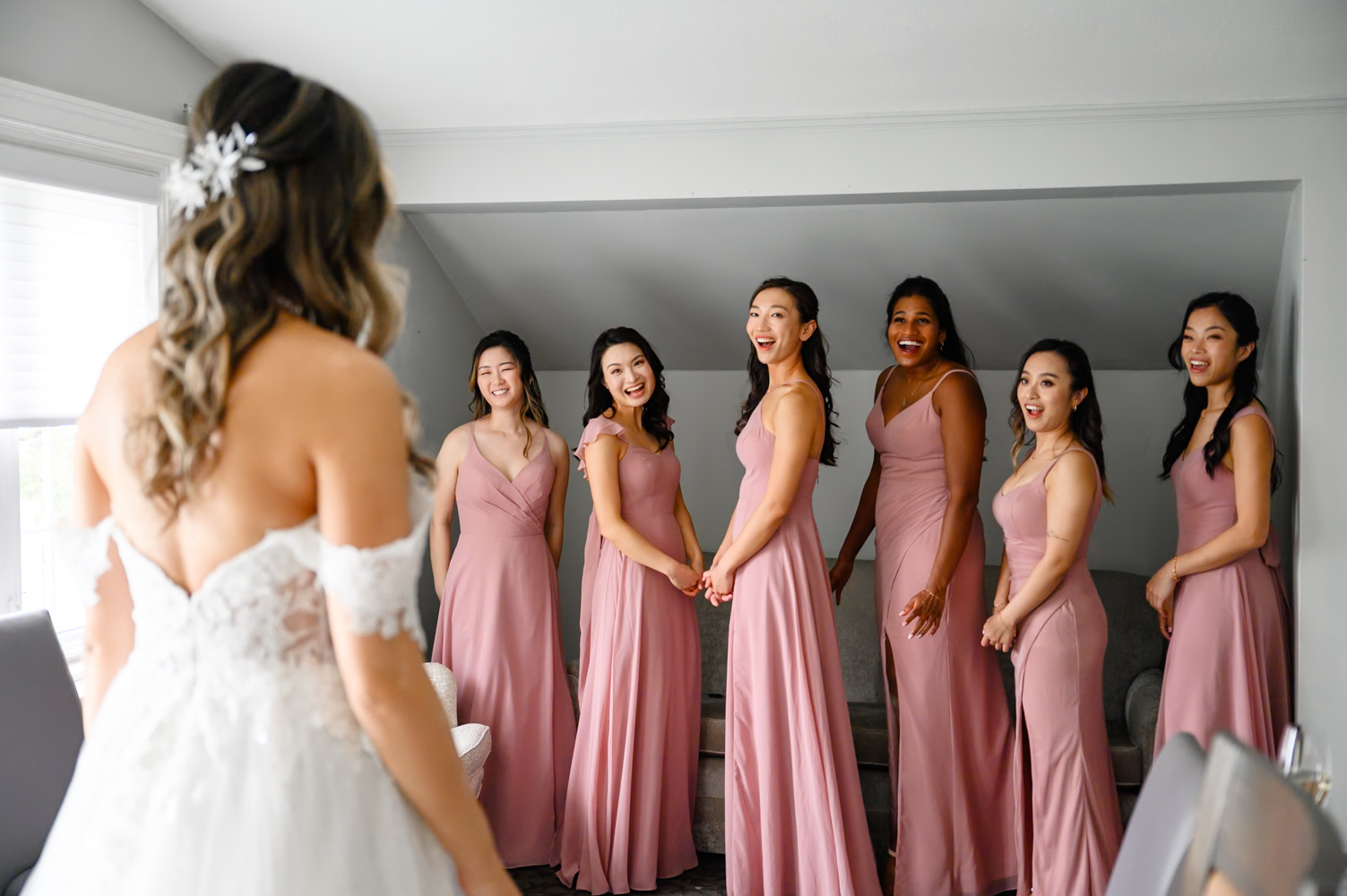 At A Toronto Hunt Golf Club wedding A bride in a white dress faces six bridesmaids wearing pink dresses, who are standing in a room looking at her and smiling.