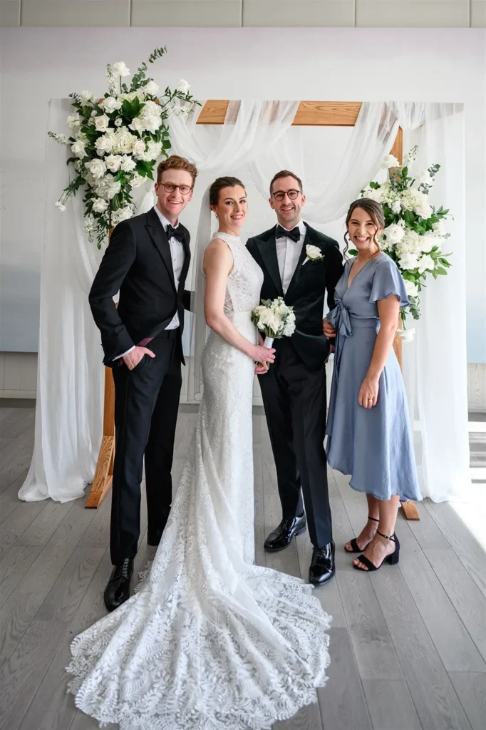 Four people pose in front of a floral wedding arch at Pearle Hotel Weddings; a bride in a lace gown and groom in a tuxedo stand together, flanked by another man in a tux and a woman in a blue dress.