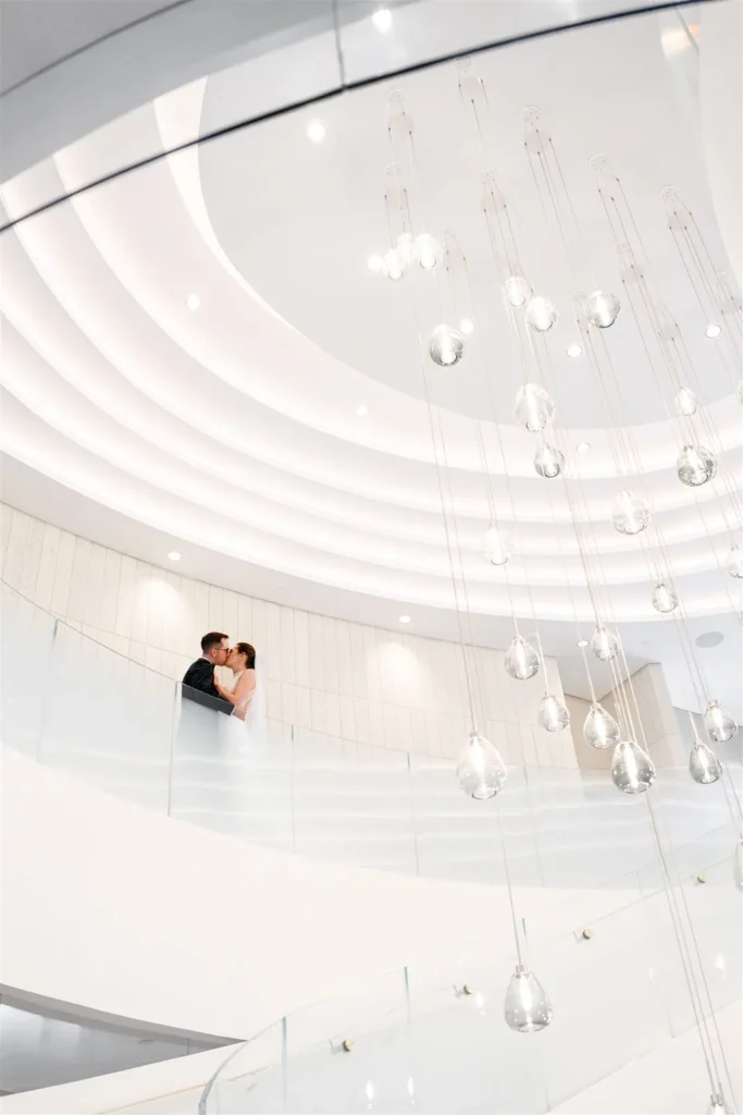 A couple stands close together on a curved balcony at Pearle Hotel Weddings, surrounded by a modern white interior with hanging glass light fixtures in the foreground.
