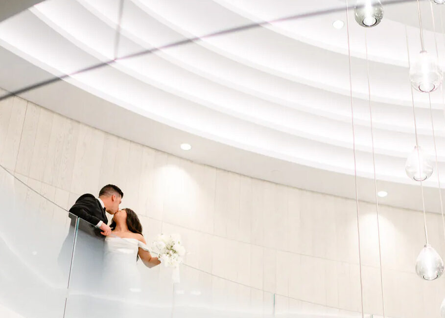 A bride and groom stand on a curved, modern balcony under circular ceiling lights, sharing an intimate moment—captured beautifully by Pearle Hotel & Spa Wedding Photography and Videography in Burlington.