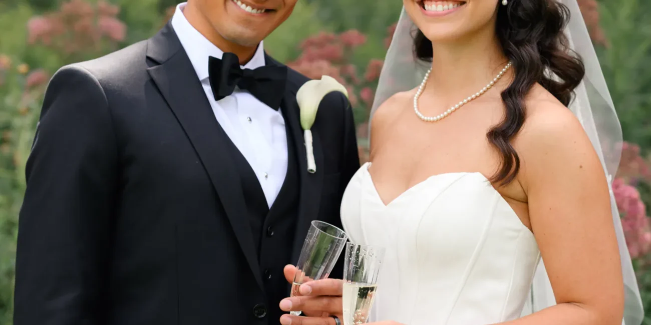 A bride and groom stand outdoors at Paletta Mansion in formal wedding attire, smiling and holding champagne glasses, with lush greenery in the background.