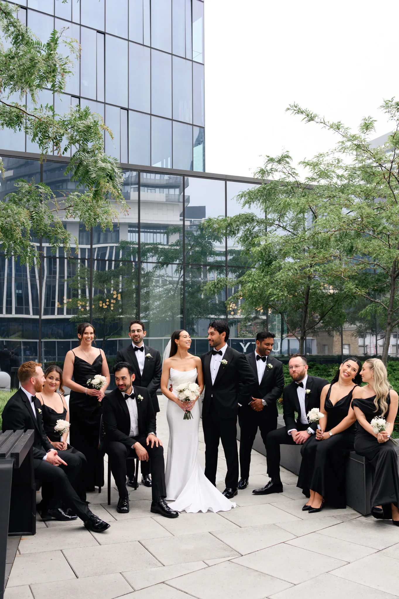 A wedding party in formal attire poses outdoors at The Quay Wedding, in front of a modern glass building, with the bride and groom standing at the center holding bouquets.