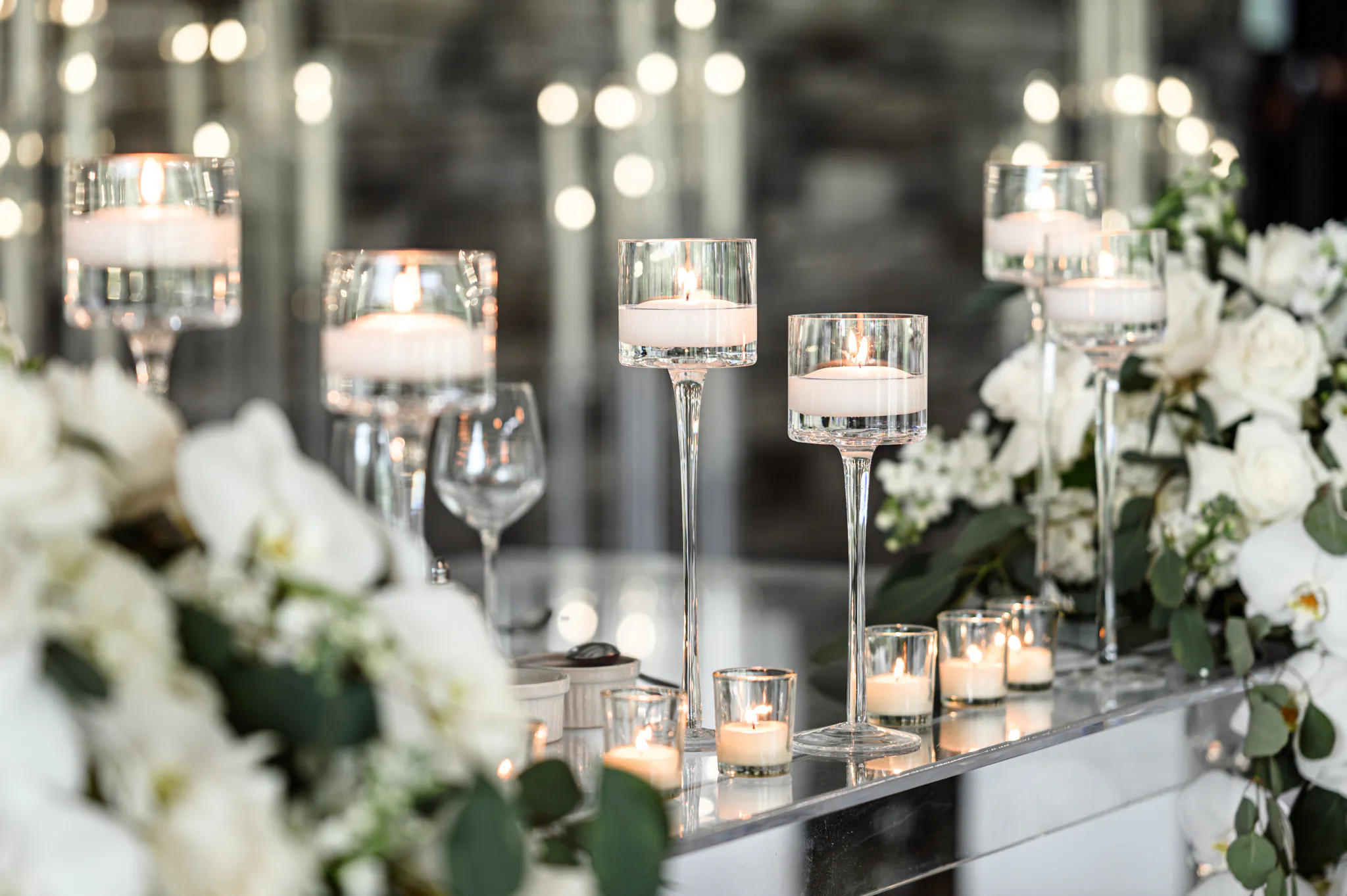 Glass candle holders with lit candles are arranged on a reflective surface, surrounded by white flowers and greenery, creating an elegant decorative display perfect for a Three Feathers Terrace Wedding.