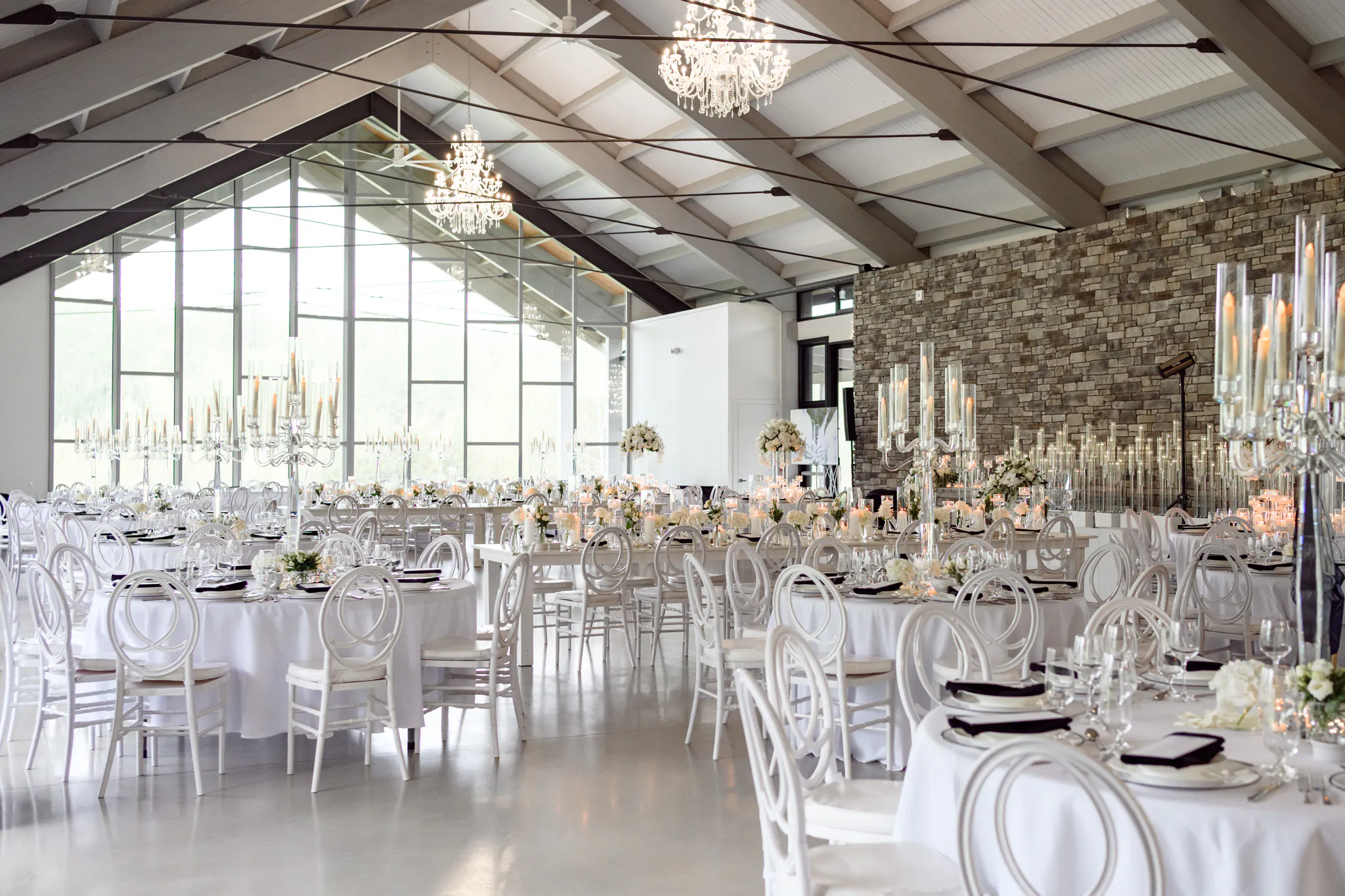 Elegant banquet hall set for a Three Feathers Terrace Wedding, with round tables, white chairs, floral centerpieces, and tall candle holders, featuring a stone accent wall and large windows.