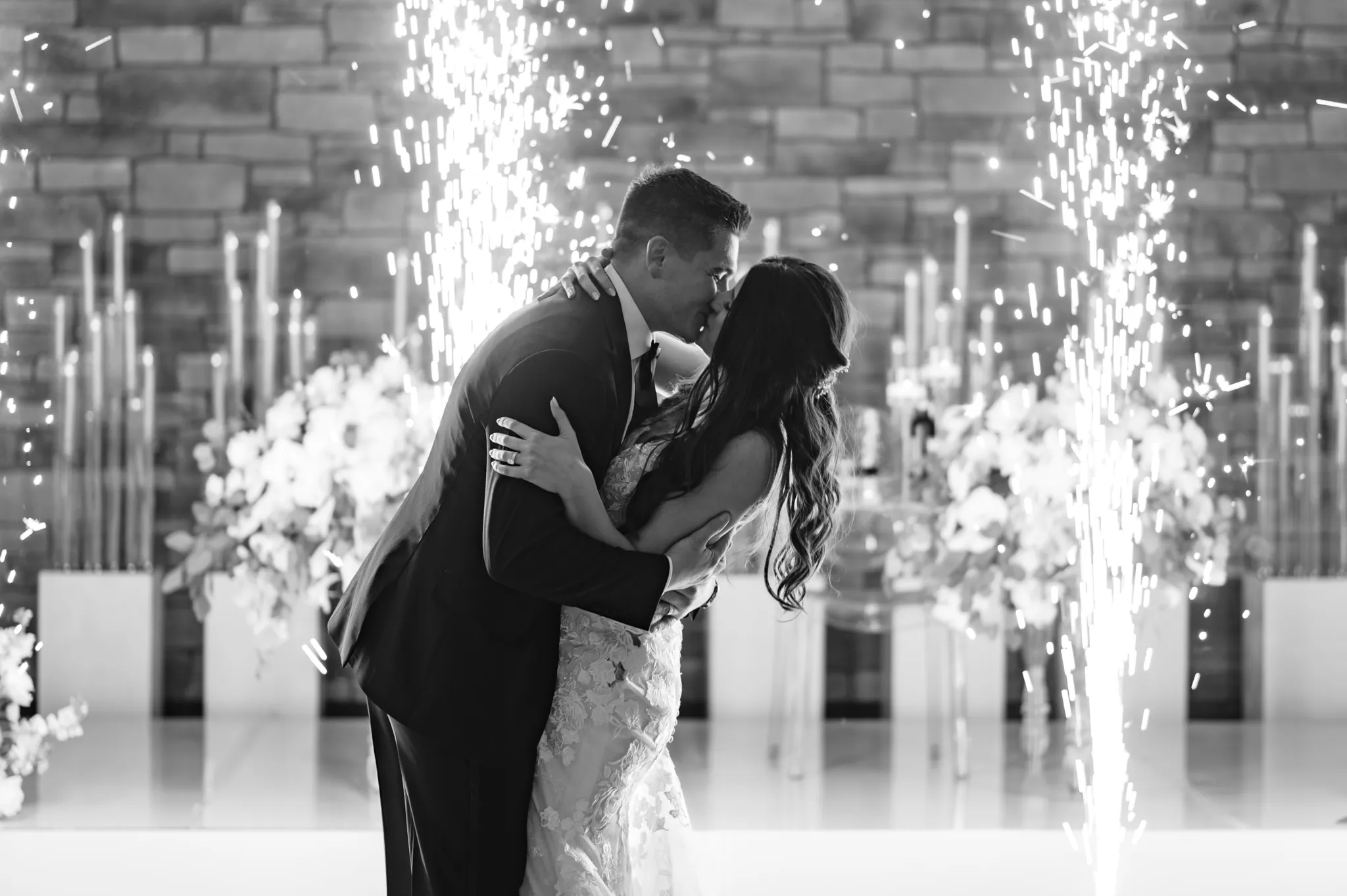 A couple dressed in formal attire embraces and kisses in front of sparklers and floral arrangements during their romantic Three Feathers Terrace Wedding ceremony.