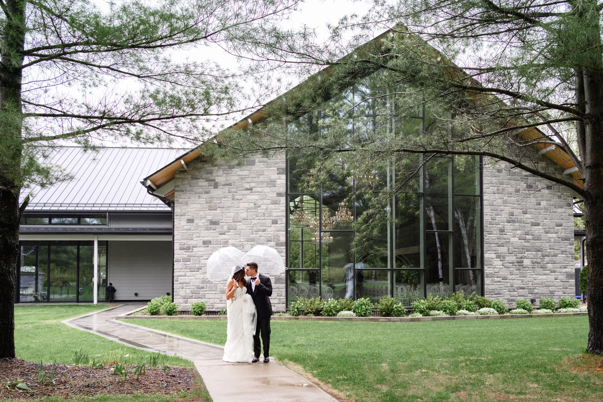 A bride and groom stand under clear umbrellas on a wet sidewalk in front of a modern stone building surrounded by greenery, capturing the romance of their Three Feathers Terrace Wedding.