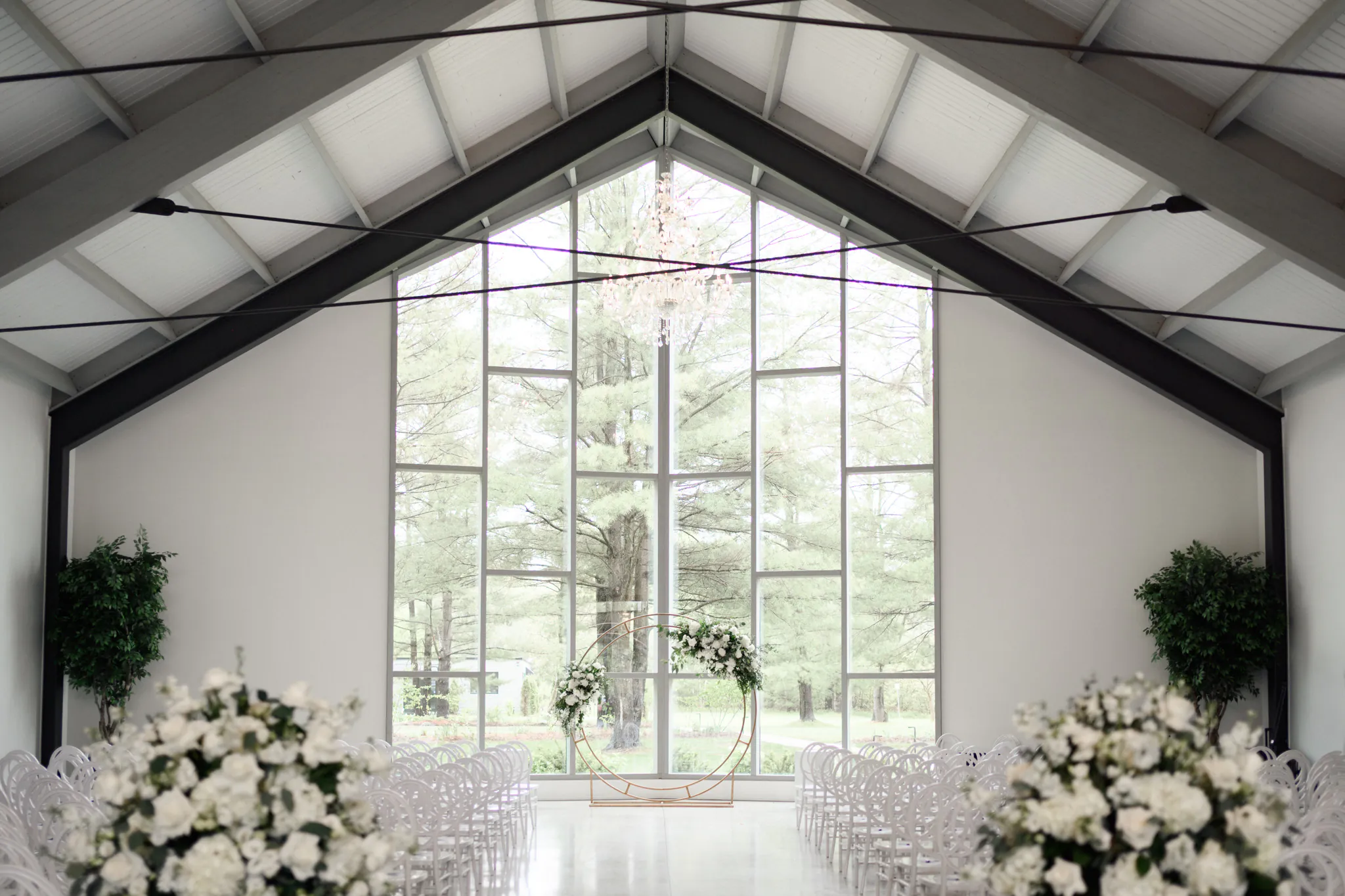 Modern Three Feathers Terrace Wedding ceremony venue with large windows, white chairs in rows, elegant floral arrangements, and lush greenery outside visible through the glass.