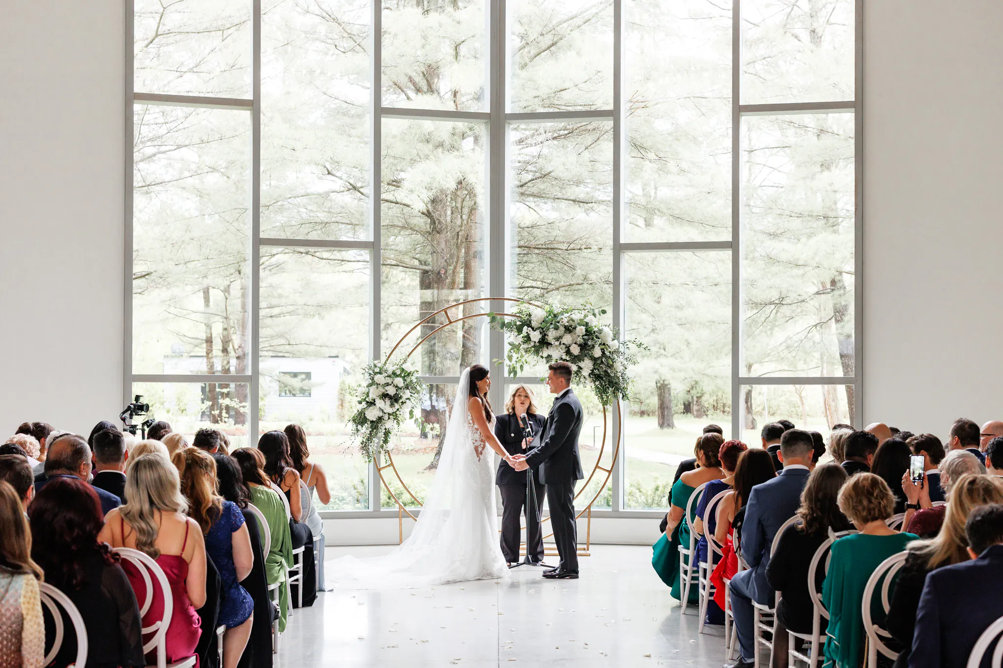 A bride and groom stand at the altar holding hands during a Three Feathers Terrace Wedding in a bright room with large windows and seated guests.