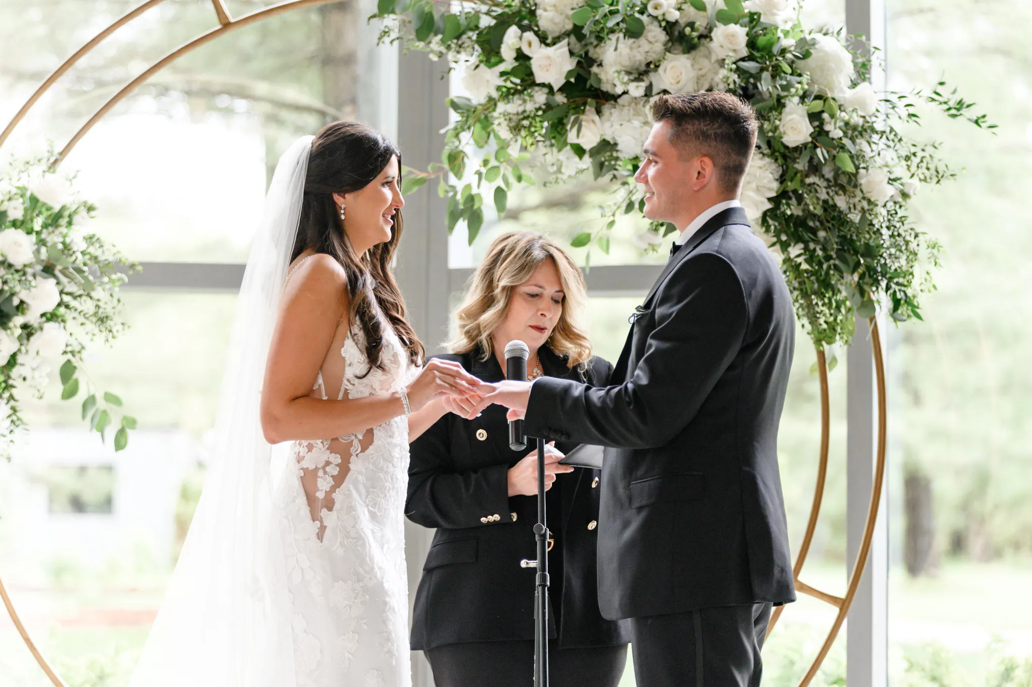 A bride and groom exchange rings during their Three Feathers Terrace Wedding ceremony, with an officiant standing between them in front of a beautiful floral arch.