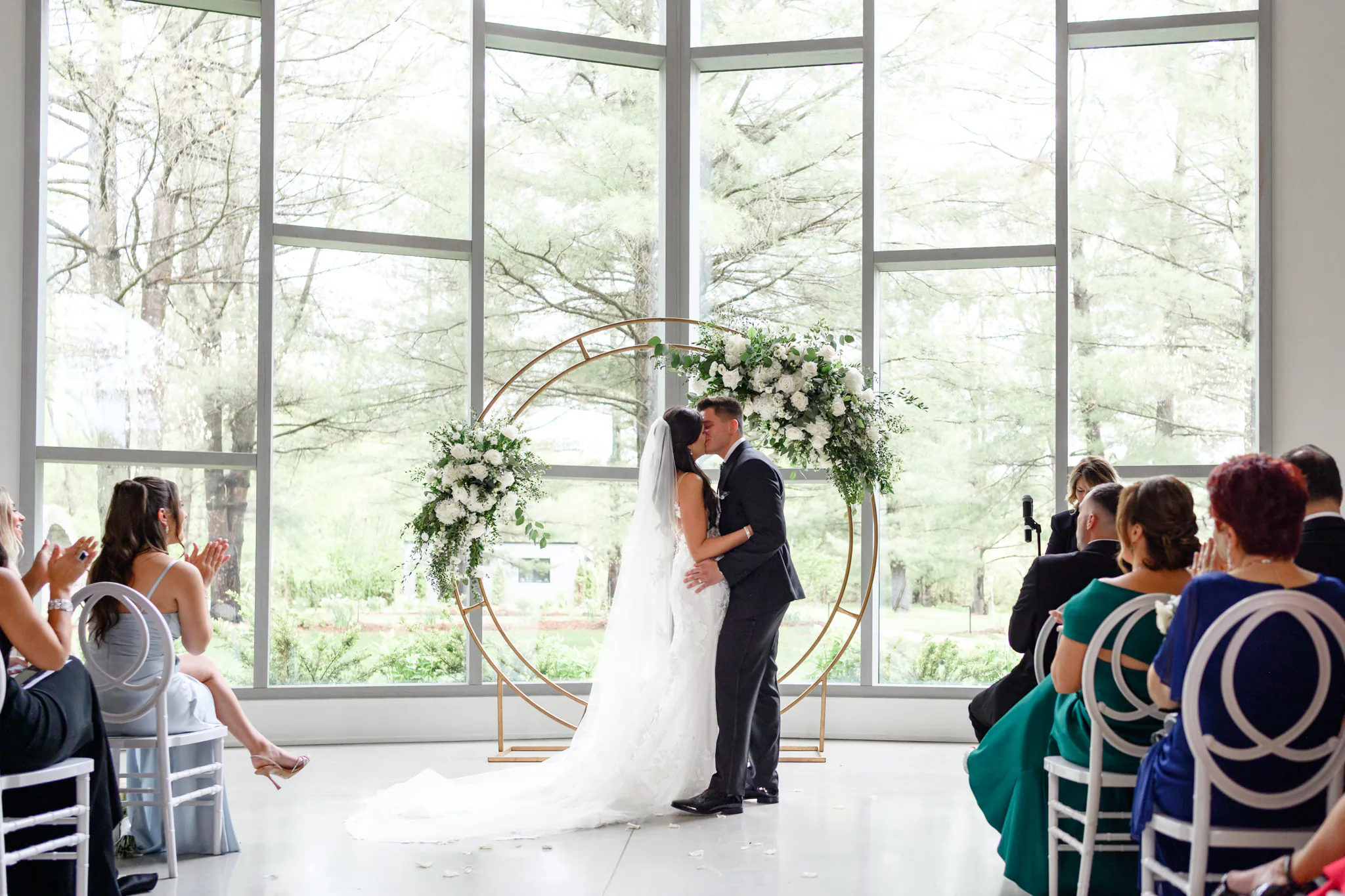 A bride and groom share a kiss at the altar during their Three Feathers Terrace Wedding, surrounded by guests seated in front of large windows and elegant floral decorations.