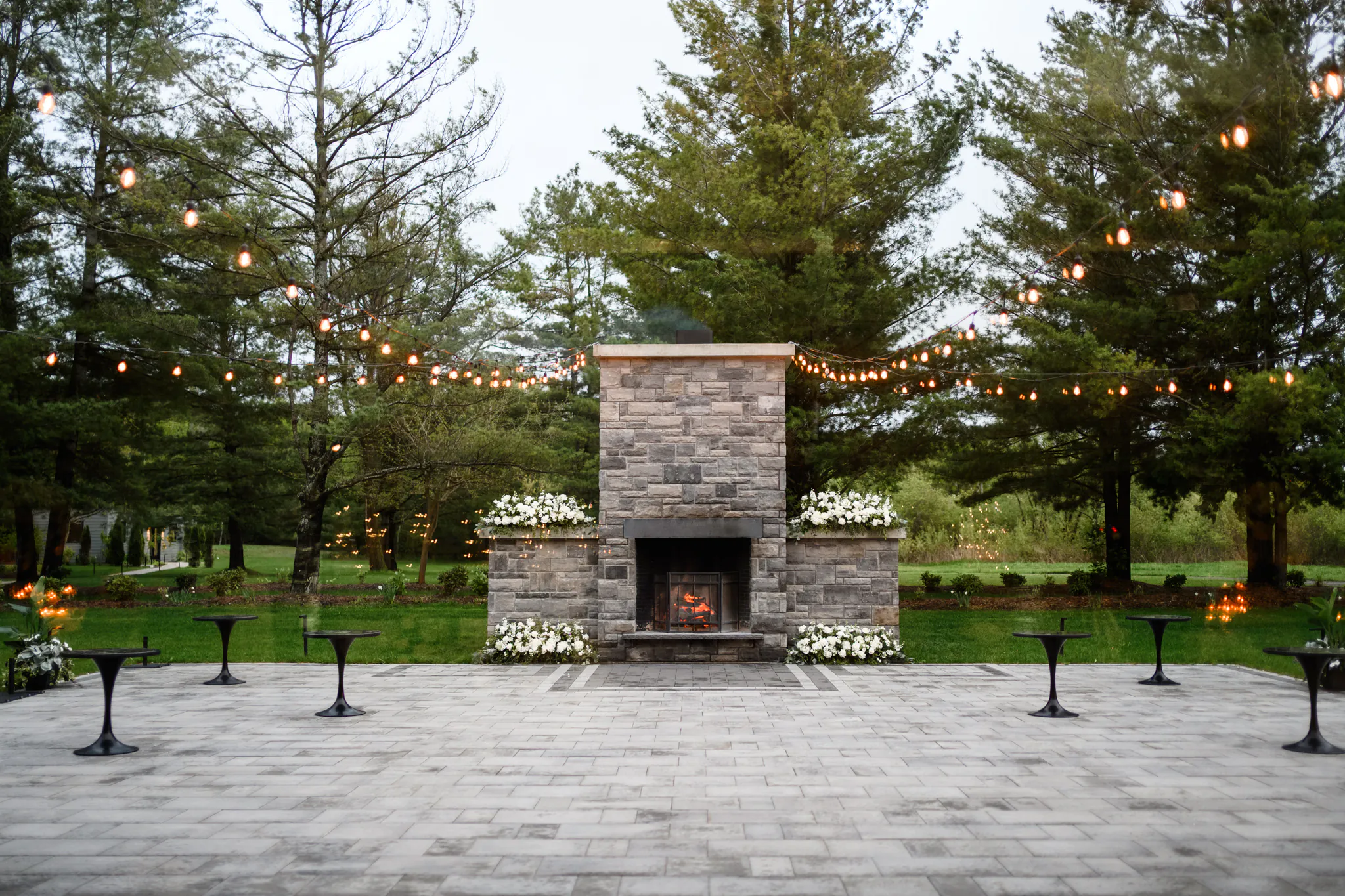 Outdoor patio with a stone fireplace, string lights overhead, small tables, and surrounding trees and greenery—perfect for a romantic Three Feathers Terrace Wedding.