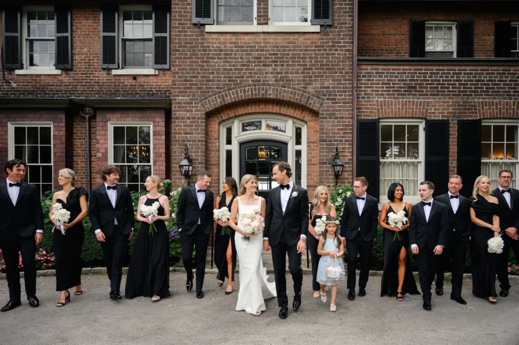 A wedding party stands in front of a brick house near the Toronto Golf Club, with the bride and groom in the center surrounded by bridesmaids and groomsmen dressed in black formal attire.