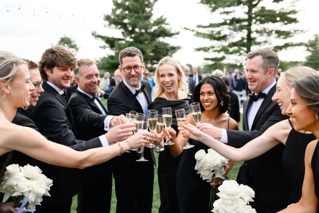 A group of people dressed in formal attire stand outdoors at Toronto Golf Club, smiling and raising glasses in a toast, with bouquets of white flowers visible.