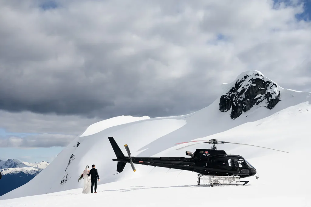 A couple in wedding attire stands on a snowy mountain next to a black helicopter, with snow-covered peaks and a cloudy sky in the background.