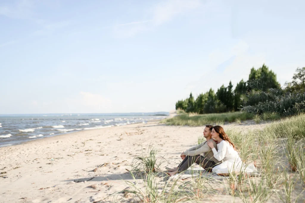 Two people sit closely together on a sandy beach near the water, surrounded by grass and trees, under a clear sky—capturing a moment that’s perfect for a memorable wedding photography experience.