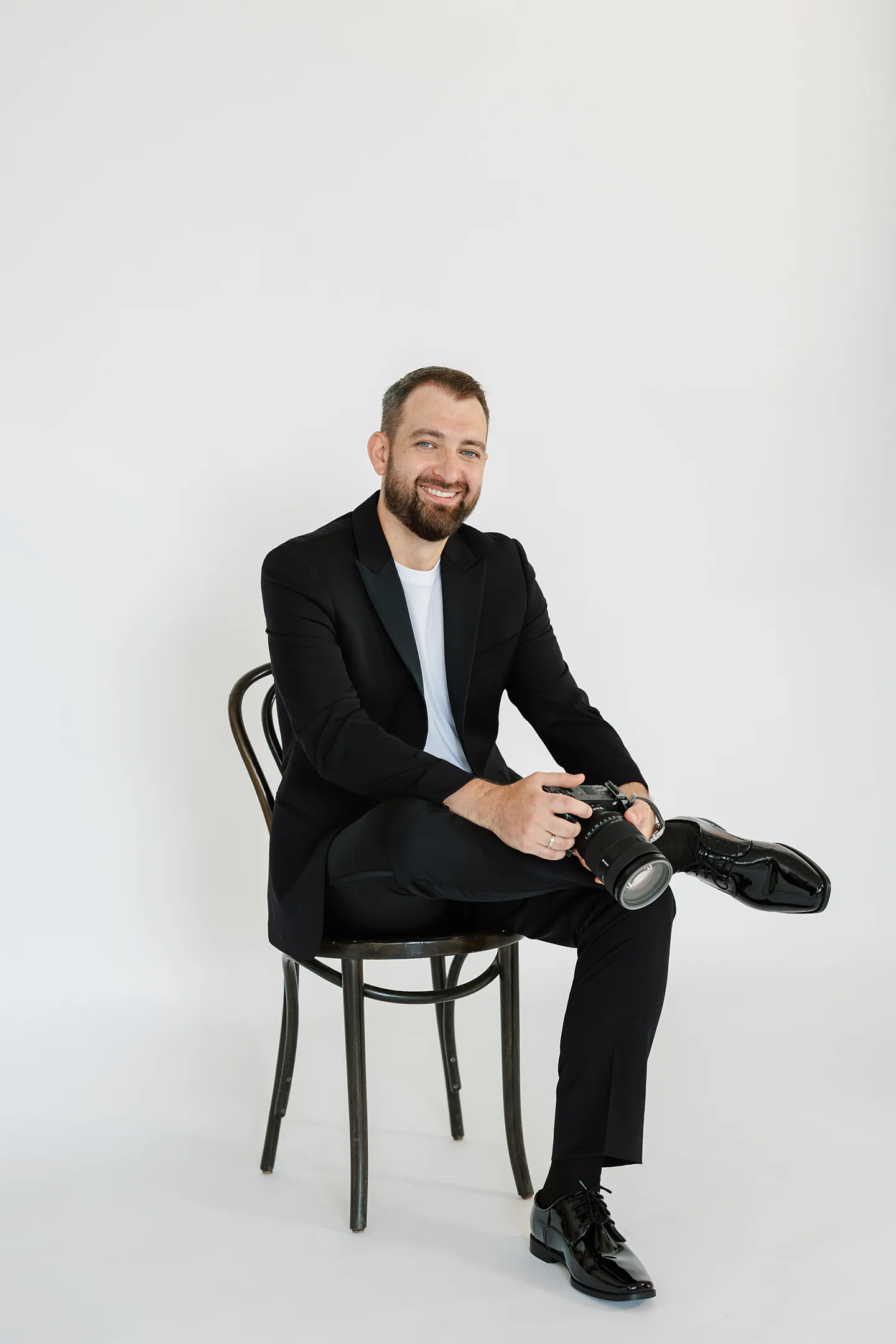 A man in a black suit sits on a chair, holding a camera and smiling, with one leg crossed over the other against a plain white background—a classic pose often seen among professional Toronto wedding photographers.